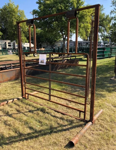 A weathered metal cattle chute stands in a grassy area, with a poster attached, surrounded by green structures and trees.