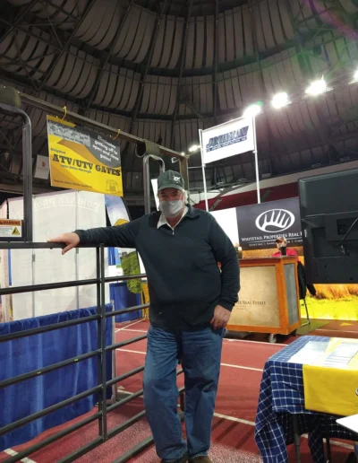 A man stands beside a metal gate at an indoor trade show, surrounded by various booths and banners highlighting outdoor products.